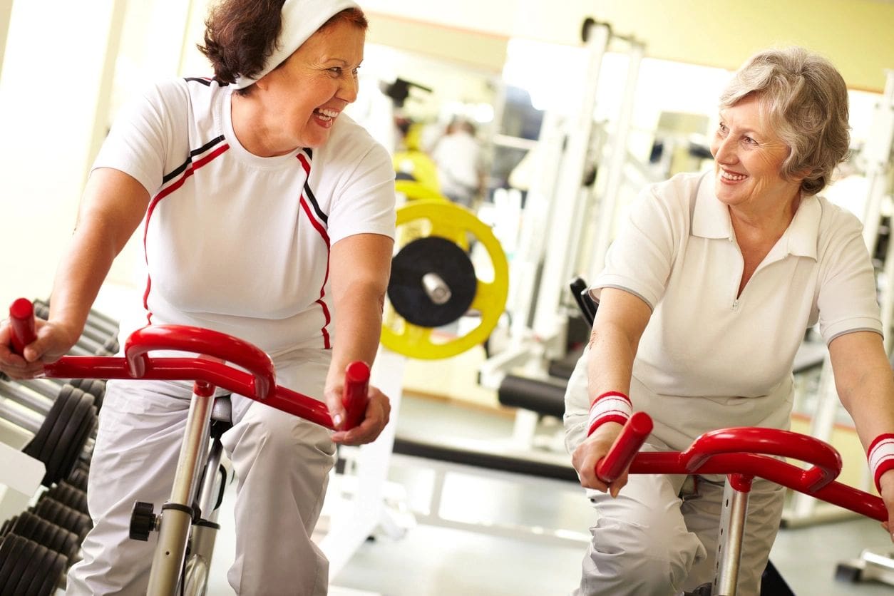 Two women are smiling while riding stationary bikes.